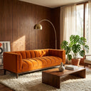 Mia 90-inch mid-century modern orange velvet channel-tufted sofa in a styled living room with wood paneling, a brass arc floor lamp, a wood coffee table, and a potted monstera plant.