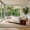 Mia 90-inch mid-century modern white boucle sofa with channel tufting and wooden legs, styled in a bright living room on a textured rug next to a walnut side table, with a potted fiddle-leaf fig plant and floor-to-ceiling windows showing tropical greenery.