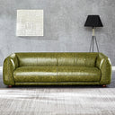 Modern olive-green distressed leather sofa with rounded armrests and low wooden feet, set against a textured gray wall with white woven square wall art, a black tripod floor lamp, and a light gray patterned rug in the foreground.
