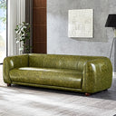 Modern olive-green distressed genuine leather sofa with curved low-profile armrests, vertical seam stitching, and round dark wooden feet, set in a contemporary living room with a gray textured wall, white wall art, a floor lamp, and a potted plant.

