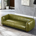 Modern olive-green distressed leather sofa with a curved low-profile backrest, wide rounded armrests, and short dark wooden feet, set in a minimalist room with gray walls, textured white wall art, a black floor lamp, and a potted plant.
