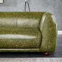 Close-up of the right side of an olive-green distressed genuine leather sofa featuring a thick bench cushion, curved armrest, visible seam detailing, and a low round dark wooden bun foot against a gray wall.
