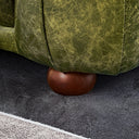 Close-up of the bottom corner of an olive green distressed genuine leather sofa with vertical channel seams, supported by a low, rounded dark brown wooden bun foot resting on a dark gray rug.
