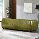 Back view of a distressed olive green genuine leather sofa with rounded edges, vertical seam detailing, and low wooden feet in a modern living room with a gray textured wall, white geometric wall art, a black floor lamp, and a potted plant.
