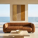 Mid-century modern brown genuine top-grain leather sofa in a minimalist living room overlooking the ocean, featuring a large vertical abstract painting on the wall and a travertine coffee table in the foreground.