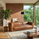Mid-century modern brown genuine top-grain leather sofa in a bright living room featuring an exposed brick wall, a large fiddle-leaf fig plant, and a tall black arched floor lamp next to a window.