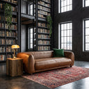 Mid-century modern brown genuine top-grain leather sofa with a green velvet throw pillow, placed on a red patterned rug in a dark industrial-style home library with tall black bookshelves and large grid windows.
