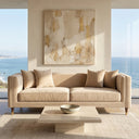 Nina 90 mid-century modern (MCM) beige velvet sofa with matching throw pillows, styled in a luxury coastal living room featuring a travertine coffee table, textured rug, and large gold abstract wall art with ocean views in the background.