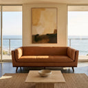 Phoenix cognac leather sofa in a modern coastal living room with ocean views, styled with a travertine coffee table, woven rug, and large abstract painting.