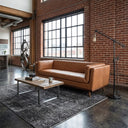 Mid-century modern Phoenix cognac leather sofa in an industrial loft living room featuring exposed brick walls, large windows, a rustic wood and metal coffee table on a grey patterned rug, and an industrial floor lamp.