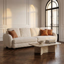 Modern living room featuring the Pina 115 cream boucle sofa with three terracotta accent pillows, styled next to a travertine stone coffee table on a dark herringbone wood floor under warm sunlight from an arched window.