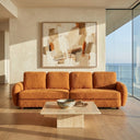 Modern Pina 115 orange boucle sofa in a minimalist coastal living room, featuring a travertine coffee table, abstract art piece, and floor-to-ceiling windows overlooking the ocean.