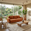 Modern living room featuring a Pina 115 orange boucle sofa placed in front of floor-to-ceiling windows overlooking a lush green garden. The interior is decorated with a textured cream area rug, a low light-wood coffee table, a woven accent chair with a draped throw, and a potted fiddle-leaf fig tree in the corner.