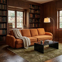 Modern Pina 115 three-seater sofa upholstered in textured burnt orange boucle fabric, styled in a cozy home library with wood-paneled bookshelves. The sofa features a chunky cream knit throw blanket, positioned on an olive green patterned rug alongside a black marble coffee table and a brass arched floor lamp.
