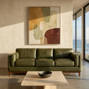 Rita 91-inch olive green top-grain leather sofa with a wooden base, positioned in a modern minimalist living room featuring a travertine coffee table, abstract wall art, and scenic ocean views through a large window.