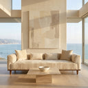 Modern Rosa 103 cream velvet three-seater sofa with wood legs in a minimalist living room featuring a travertine coffee table, abstract art, and ocean views.