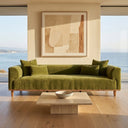 The Rosa 103 green velvet sofa with matching throw pillows, styled in a minimalist modern living room with a low travertine coffee table, large abstract art piece, light wood floors, and floor-to-ceiling windows overlooking a sunny ocean view.