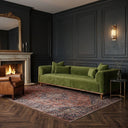A Rosa 103 green velvet sofa with matching pillows in a luxury living room with dark paneled walls, a burning fireplace under a gold-framed mirror, a brown leather armchair, and an oriental rug on herringbone hardwood floors.