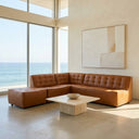 Selia 92-inch comfy chic modular sectional sofa and matching ottoman in cognac brown tufted leather, styled in a bright modern living room with floor-to-ceiling ocean view windows and a low stone coffee table.