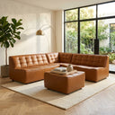 Selia 92-inch comfy chic modular L-shaped sectional sofa with matching square ottoman in cognac brown tufted leather, styled on a neutral area rug in a sunlit living room with floor-to-ceiling windows.