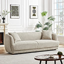 Modern cream luxury fabric sofa with rounded armrests and matching accent pillows in a bright living room with framed wall art and a potted palm plant.
