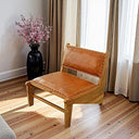 A low-profile accent chair with a natural wood frame and tan leather seat and backrest, accented by studded nailhead trim along the edges. The chair sits on a hardwood floor bathed in natural sunlight beside a tall black vase filled with blooming pink branches, positioned in front of a window with beige and sheer curtains.