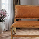 Warm tan leather accent chair bench with a natural wood frame, featuring an upholstered backrest and seat lined with decorative nailhead trim, placed on light wood flooring beside a white vase with purple flowers.