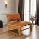 Rustic low-profile accent chair with cognac-tan leather upholstery and decorative nailhead trim along the borders of the seat and backrest. The chair has a sturdy natural light wood frame with block legs, positioned on a wooden floor in an interior beside tall curtained glass doors and a vase of pink flowers.