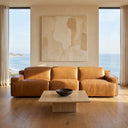 Valeria 120 triple power tan leather recliner sofa in a luxury modern living room with floor-to-ceiling windows overlooking the ocean. A travertine coffee table sits in front, and a textured abstract painting hangs on the wall behind.