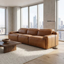 Valeria 120 triple power tan leather recliner sofa in a modern high-rise living room with floor-to-ceiling windows showing a city skyline view, featuring a wooden coffee table and a minimalist black floor lamp.