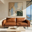 Vanessa 89-inch tan genuine top-grain leather sofa in a modern living room with a large abstract painting, travertine coffee table, and a floor-to-ceiling window showing a coastal ocean view.