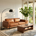 Vanessa 89-inch tan genuine top-grain leather sofa in a mid-century modern living room setup with a matching wooden coffee table, a black arched floor lamp, a large potted fiddle-leaf fig plant, and a patterned cream area rug against a white brick wall filled with warm natural sunlight.