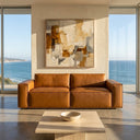 Vanna 93-inch mid-century modern cognac vegan eco-leather sofa in a sunlit living room with floor-to-ceiling ocean view windows and abstract wall art.