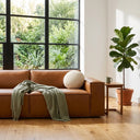 Vanna 93-inch Mid-Century Modern cognac brown vegan eco-leather sofa styled with a sage green throw blanket and round cream bouclé pillow in a sunlit living room with large windows and a potted fiddle-leaf fig plant.